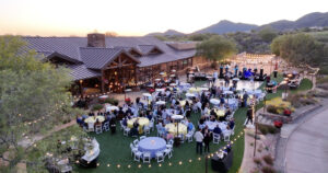 tables and chairs outside at dusk underneath string lights as people sit and eat listen to a concert
