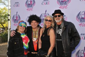 a group of four people smiling in rock n roll outfits in front of a white banner at care's rock for hope bash for cancer research at desert mountain club