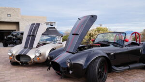 two 1965 cobras, a light beige and a black one with red stripes down the middle, sitting in a driveway under overcast skies at desert mountain club, two of the many classic cars in arizona