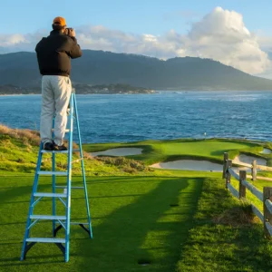 evan schiller, a premier golf photographer, standing on a green ladder on a tee box at pebble beach taking a photo
