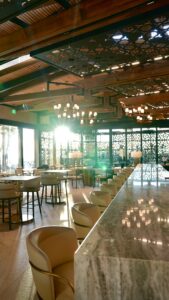 Interior shot of the bar area at Renegade Clubhouse at Desert Mountain Club, winner of a Gold Nugget Award. Sunshine peaks through decorated metal shades and lights up a marble bar top underneath large wooden beams supporting large sky lights and designer lights
