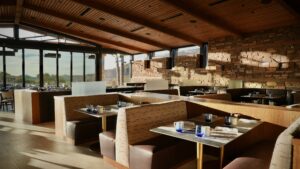 Interior of the dining room at Renegade Clubhouse at Desert Mountain Club, winner of a Gold Nugget Award, showing natural light flowing through floor to ceiling windows, large wooden beams and a stone wall interior to highlight its modern rustic design